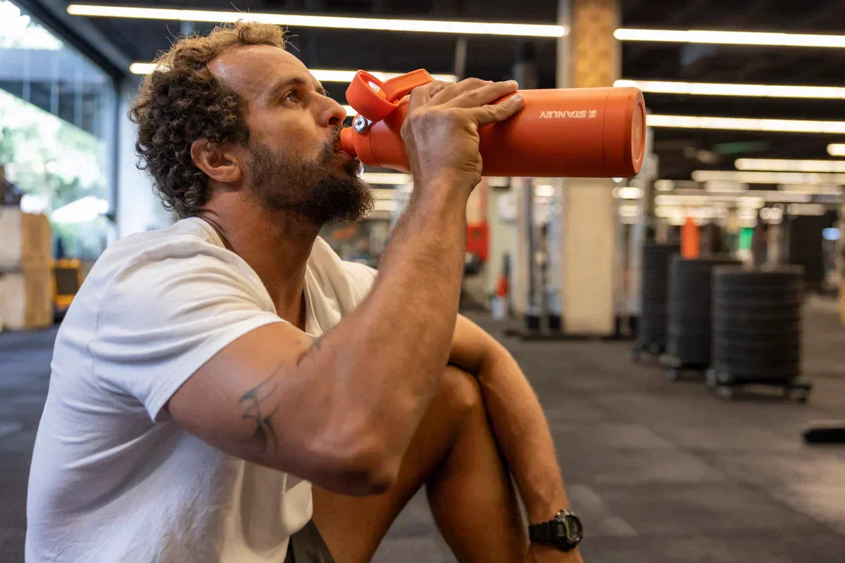 Um homem sentado usando a garrafa Stanley para se hidratar durante os exercícios físicos na academia.