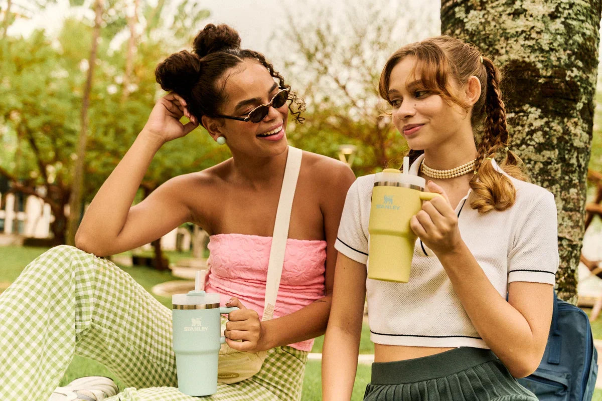 Duas amigas sentadas lado a lado, cada uma segurando o seu Mini Quencher Stanley.