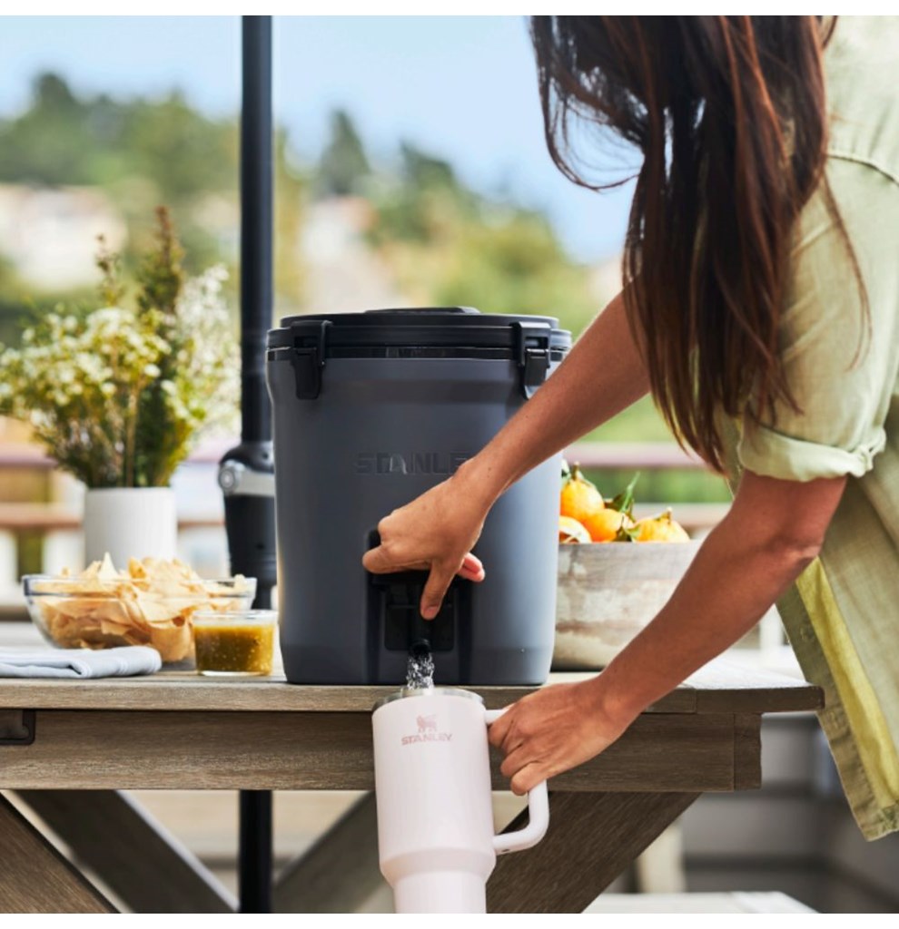 Uma mulher está enchendo um copo Quencher na cor branca com líquido vindo do Jug térmico, um grande recipiente cinza escuro que está sobre uma mesa de madeira ao ar livre. O recipiente tem uma torneira na parte inferior. Na mesa, há também um prato com batatas chips, um pequeno pote com molho e um vaso com flores. Ao fundo, há uma tigela com frutas. A cena parece ser em um ambiente externo, com vegetação ao fundo.